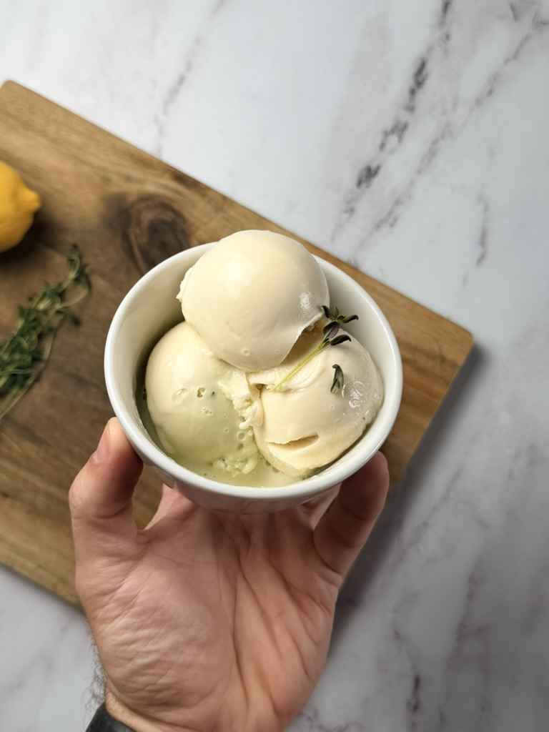 Hand-held cup of vanilla protein ice cream scoops on a marble surface with lemon and thyme